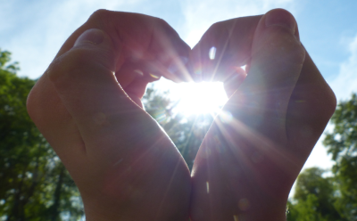 Two hands form a heart shape outdoors, with the sun in the centre of the heart and greenery seen in the background
