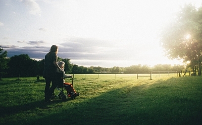 A person pushes another person sitting in a wheelchair through a beautiful green field that is lit up with sunshine