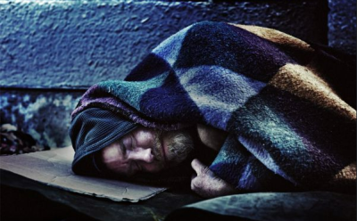 A person sleeping on a cardboard box with a jumper and a blanket wrapped closely around their head